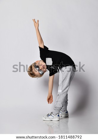 Blond kid boy in sunglasses, black t-shirt, gray pants and sneakers does gymnastics bending exercises tilts body forward arms spread wide over gray background