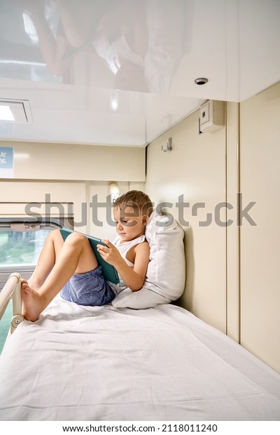 Blond\
little boy watches cartoons via tablet PC and leaning on soft\
pillow on top shelf in train compartment in\
morning