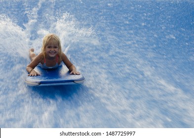 Blond Little Girl On The Surf Board Floating Against The Water Flow. Flow Rider Sport. Girl Swims On A Board On A Wave.  