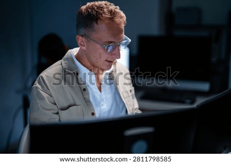 A blond male network security analyst scans lines of code lit by the screen glow inside a modern security operations centre after hours. The image conveys defending the corporate network.