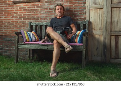 Blond Man In Black T-shirt, Green Shorts And Sandals Sits On A Bench In Backyard.