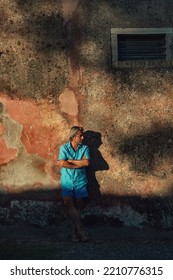 Blond Man In Blue Shirt And Swim Shorts Stands Against Weathered Pink Plaster Wall In Late Afternoon Sunlight.