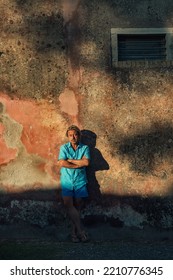 Blond Man In Blue Shirt And Swim Shorts Stands Against Weathered Pink Plaster Wall In Late Afternoon Sunlight.