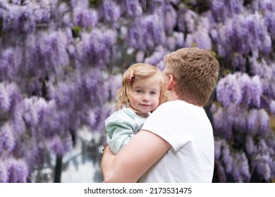 Blond Man Holding Cute Baby Girl,daughter,looking At Camera,near Glucinum Flowers,purple,very Peri Wistaria Tree.Father And Toddler,child,kid,infant Together.Happy Parenthood,lifestyle.Copyspace.