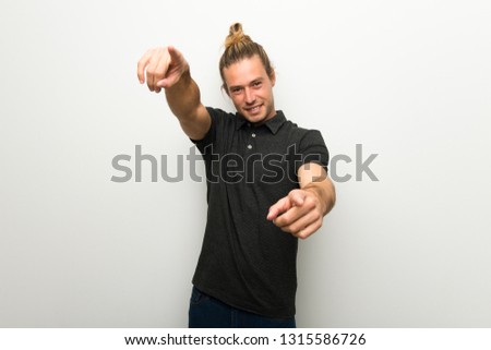 Blond man with long hair over white wall points finger at you while smiling