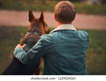 A Blond Man Sits And Hugs A German Shepherd Dog. Rear View.