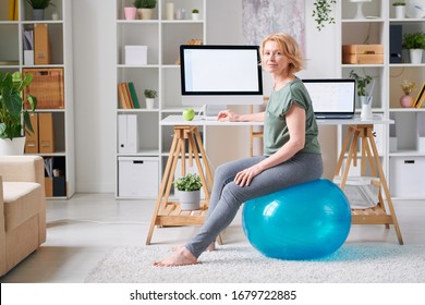 Blond mature female in activewear sitting on fitball by desk in front of computer monitor while going to exercise at home - Powered by Shutterstock