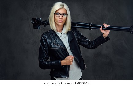 A blond photographer female dressed in a black leather jacket holds professional digital camera and a tripod.
