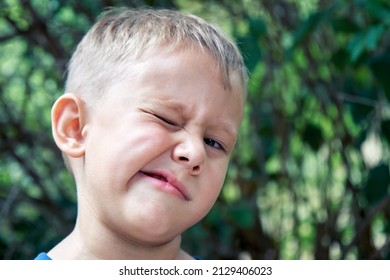 Blond Preschooler Grimaces Making Weird Face Expressions. Boy Has Fun Walking Outdoors In Shade Of Trees In Park On Summer Holiday Closeup