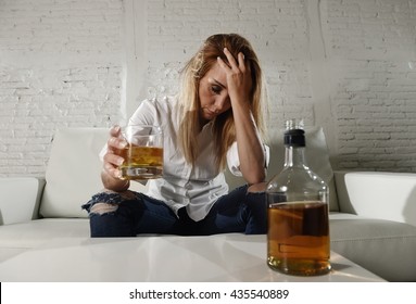 blond sad and wasted alcoholic drunk woman sitting at home sofa couch drinking scotch whiskey holding glass depressed lonely and suffering hangover in alcoholism and alcohol abuse concept