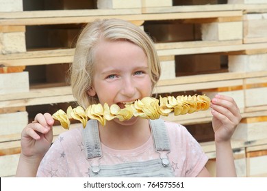 Blond School Girl Dressed In Bib Overalls Eating Potato Chips On A Stick. Junk Food, Street Food, Snack On A Walk. Country Style.