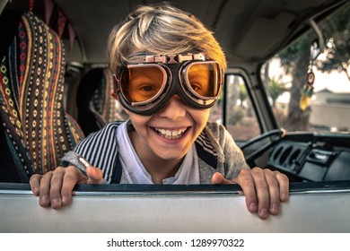 Blond smiling child looks out the window of vintage camper wearing aviator goggles sitting on colorful seat, puts his hands on the car door on the palms and sunset background. Vintage funny portrait - Powered by Shutterstock