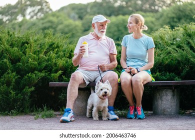 A Blond Sporty Daughter Take Care Of Her Father 80 Year. Senior Man And Young Adult Woman Very Happy Sitting On Bench At Summer Park With Is Dog. Concept Care And Nurse With Old People.