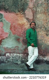 Blond Tanned Man In Green Shirt And White Pants Stands In Front Of Weathered Pink Plaster Wall.