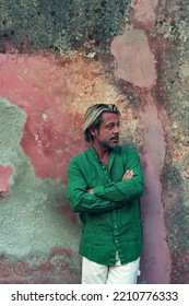 Blond Tanned Man In Green Shirt And White Pants Stands In Front Of Weathered Pink Plaster Wall.