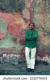 Blond Tanned Man In Green Shirt And White Pants Stands In Front Of Weathered Pink Plaster Wall.