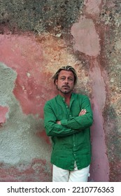 Blond Tanned Man In Green Shirt And White Pants Stands In Front Of Weathered Pink Plaster Wall.