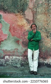 Blond Tanned Man In Green Shirt And White Pants Stands In Front Of Weathered Pink Plaster Wall.