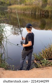 Blond Teenage Boy Wearing Black And Gray Outfit, Standing At The Edge Of The Lake Fishing