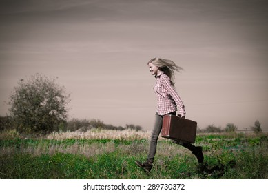 Blond teenage girl walking fast carring retro suitcase over rural background copyspace on summer cloudy day