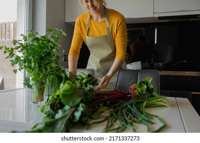 Blond Woman In Apron Cooking Healthy Fresh Vegetable Salad In The Home Kitchen. Female Holding Cucumber Plate Or Bowl For Washing. Organic Farming Dish For Loosing Weight In Summer. Learning To Cook