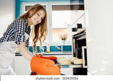 Blond woman baking a bread rolls in kitchen oven. She pulls rolls from the oven. Hands protected by orange kitchen gloves.  - Powered by Shutterstock