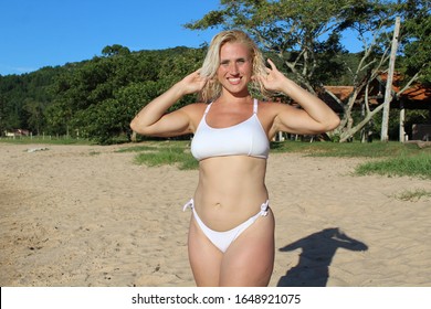 Blond woman at the beach holding her hair with sand and green trees in the background smiling at the camera in a sunny day