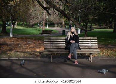 Blond Woman With Black Mask Looking At The Mobile Sitting On A Park Bench