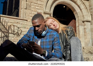 Blond Woman With Closed Eyes Leaning On Shoulder Of Black Boyfriend Using Smartphone While Sitting On Sunlit Steps Outside Old Building On Weekend Day