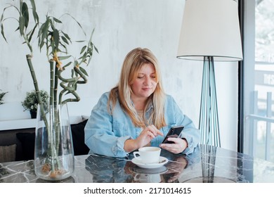 Blond Woman In Denim Shirt Sitting At Table Typing Text Messages On Her Phone Using Modern Smartphone