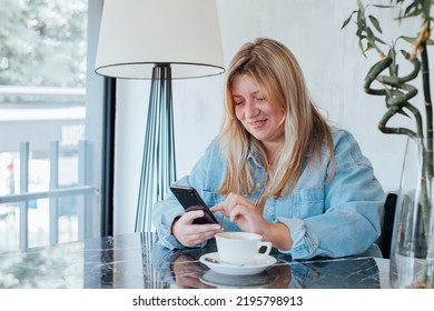 Blond Woman In Denim Shirt Sitting At Table Typing Text Messages On Her Mobile Phone