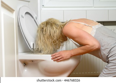 Blond woman in a grey dress kneeling down vomiting into a toilet conceptual of an illness or drunkenness after a party