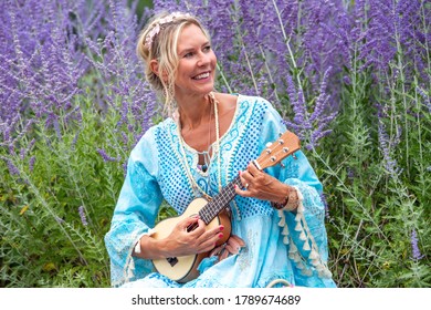 Blond Woman In Her 40s Sitting At Field Of Flowers And Playing The Guitar