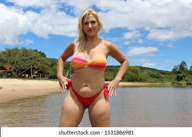 Blond woman with her hands on her hips wearing a bikini at the beach with water, sand, blue sky and green trees in the background