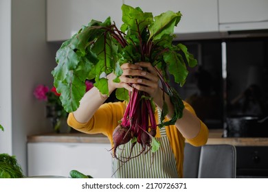Blond woman hiding face behind bunch of beetroots with leaves, cook fresh organic salad in kitchen on summer weekend. Eco friendly food, healthy eating and diet at home. Joying farming vegetables