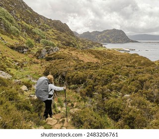 Blond woman with hiking poles and big hiking backpack looking at a lush coastal landscape near Glenuig, in the west coast of Scotland - Powered by Shutterstock