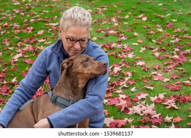 Blond Woman Hugging Doberman Mix Dog, Outside On Green Lawn Covered In Red Fall Leaves
