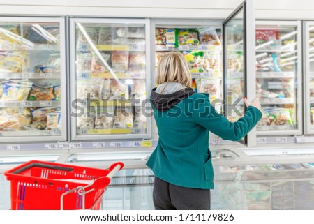 A blond woman picking frozen goods from a supermarket refridgerator with a red shopping cart next to her. Casual shopping. 