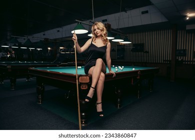 Blond woman playing enjoying billiard, hold cue, white billiard balls on table with green surface in billiard club. Pool game snooker pyramid player

