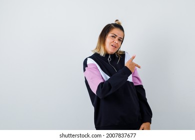  Blond Woman Pointing Down, Standing Sideways In Black Tracksuit And Looking Thoughtful.