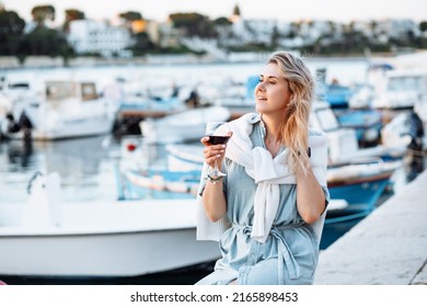 Blond woman with shirt around neck, holding tasty wineglass on vacation in travelling near boat port. Enjoy sunset view