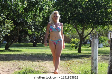 Blond woman walking on the beach trail in the sand on a sunnyday with grass and green trees in the background wearing a blue bikini