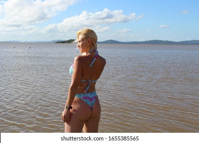 Blond woman in the water at the beach staring at the far wit the water and blue sky with clouds in the background and mountains in the horizon, wearing a blue bikini