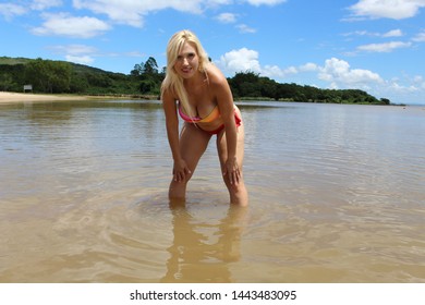 Blond woman in the water on the beach with hands on her knees withy water and sky in the background