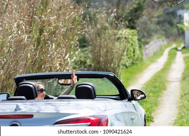 Blond Woman Waves In A Cabriolet Car While Driving Away On A Countryside