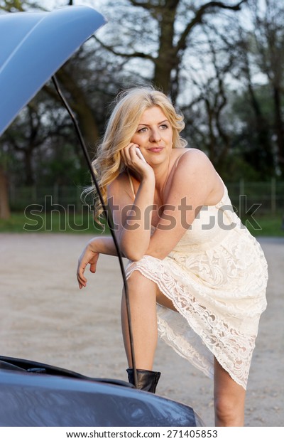 Blond woman wearing white lace dress and short\
black boots in front of the trunk of a car thinking what to do\
after car break down.