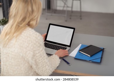 Blond Woman Working On A Laptop Computer As She Sits At Her Desk, Over The Shoulder View Of The Blank White Screen