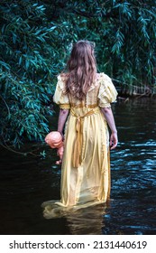 blonde Frau in gelbem historischem Kleid, die im Wasser steht und eine Puppe hält