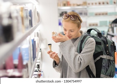 Blond young female traveler wearing coat and travel backpack choosing perfume in airport duty free store. Casual lady testing and buying cosmetics on the go in a beauty store. - Shutterstock ID 1304754997