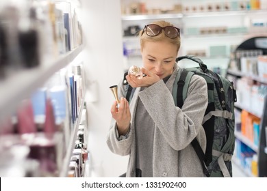 Blond young female traveler wearing coat and travel backpack choosing perfume in airport duty free store. Casual lady testing and buying cosmetics on the go in a beauty store. - Shutterstock ID 1331902400